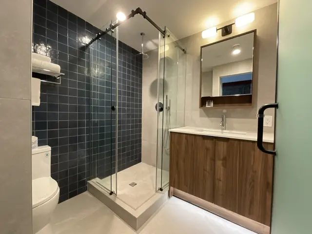 Corner glass shower with a dark tile wall beside the walnut vanity in a MODO Hotel guest bathroom