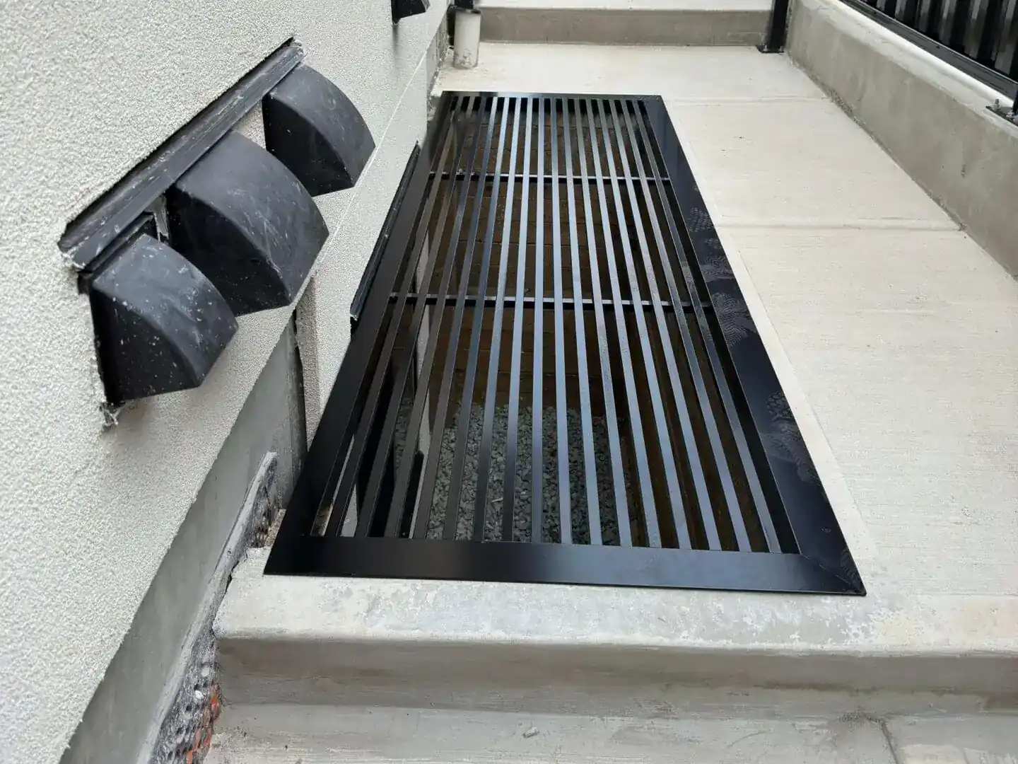 Black metal window well grate cover set into a concrete side walkway